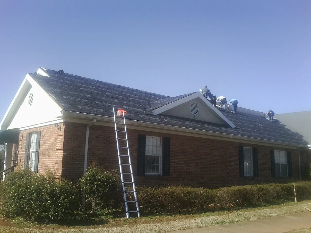 Crew tearing shingles off a brick ranch roof with a ladder set — Metal Roof Repair work by our crew in Leroy
