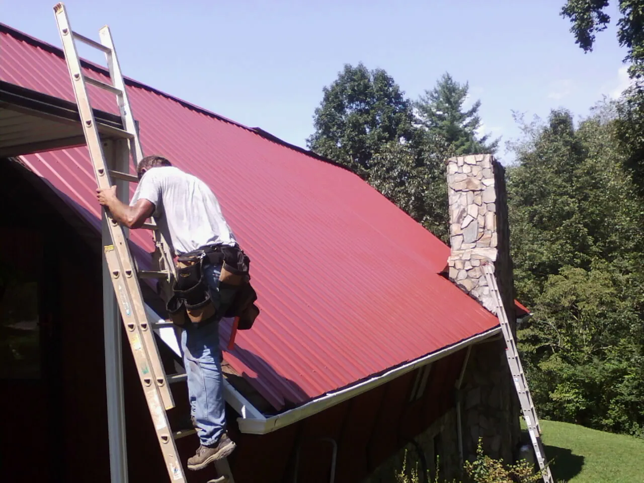 Roofer on a ladder beside a red metal roof and stone chimney — Soffit & Fascia Repair by Roofing Brothers in Leroy