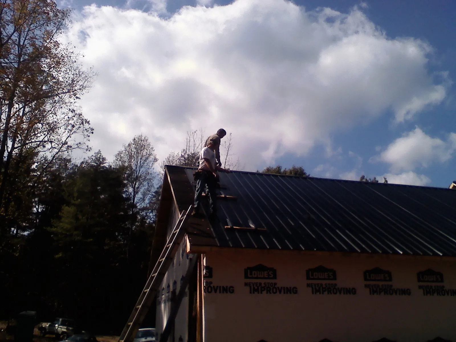 Roofer fastening dark metal panels high against a cloudy sky, from a Roof Inspection job in Leroy